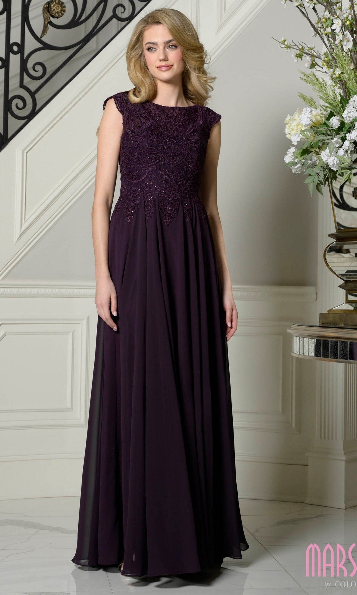 A woman stands indoors by a staircase and floral display, wearing the Marsoni M238—a sleeveless, floor-length eggplant gown with lace bodice. Perfect as a mother of the bride dress for elegant events.